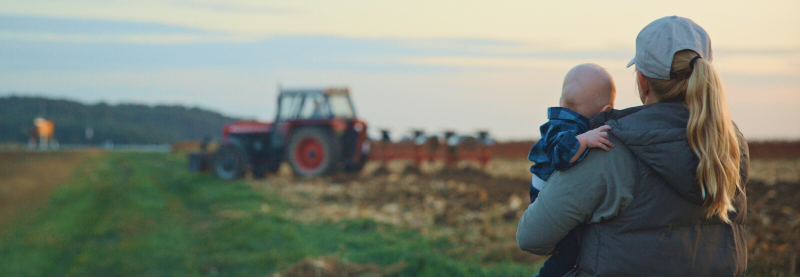 A woman holding a young baby and looking at a farm tractor in the distance